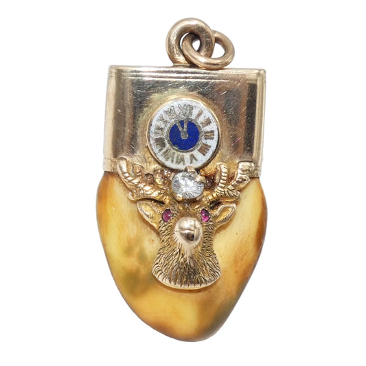An elk tooth with 10k gold featuring an elk head motif with a circular clock face at the top, accented with a blue enamel center, a clear central stone, and two pink gemstones in the eyes