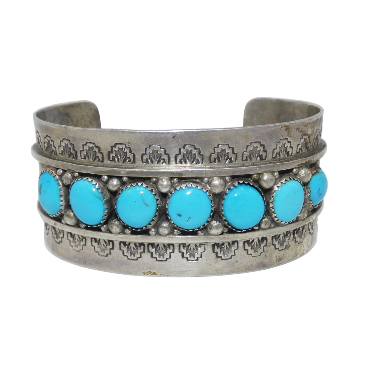 A silver cuff bracelet featuring seven round turquoise stones in a row, set with small silver beads and bordered by engraved geometric patterns